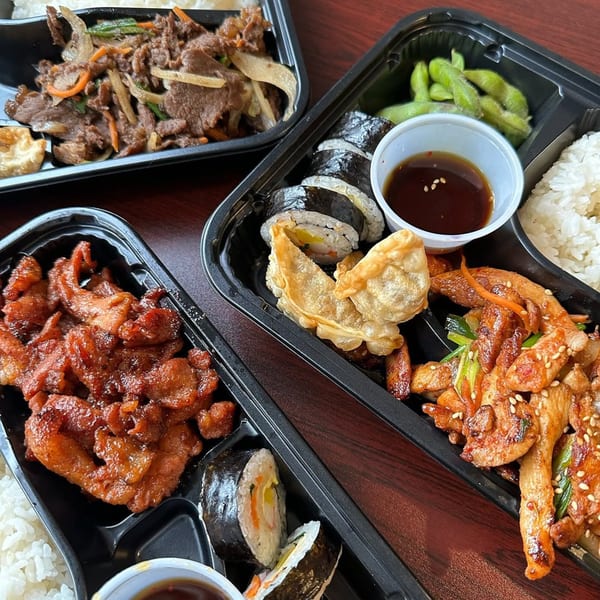 Three black takeout containers filled with various Korean dishes, including bulgogi, spicy pork, kimbap, and rice.
