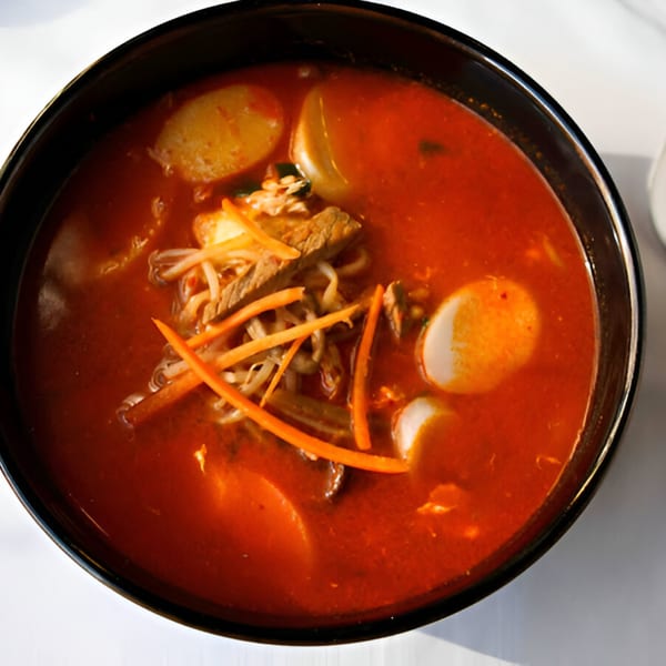 A black bowl of vibrant red Korean soup with beef, rice cakes, and vegetables.